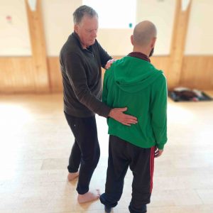2 men are standing next to one another in a movement studio with a wooden floor.. One has a hand on the lower back of the other, and one one hand on their shoulder, as they offer some listening touch.